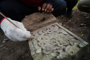 A person wearing a white glove delicately cleans an intricately carved stone artifact with a small brush, revealing floral patterns. Another person’s hand is visible above the artifact.