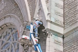 A construction worker in a hard hat stands on a lift, repairing or inspecting the ornate stone facade of a historic building with arched windows and detailed stonework.