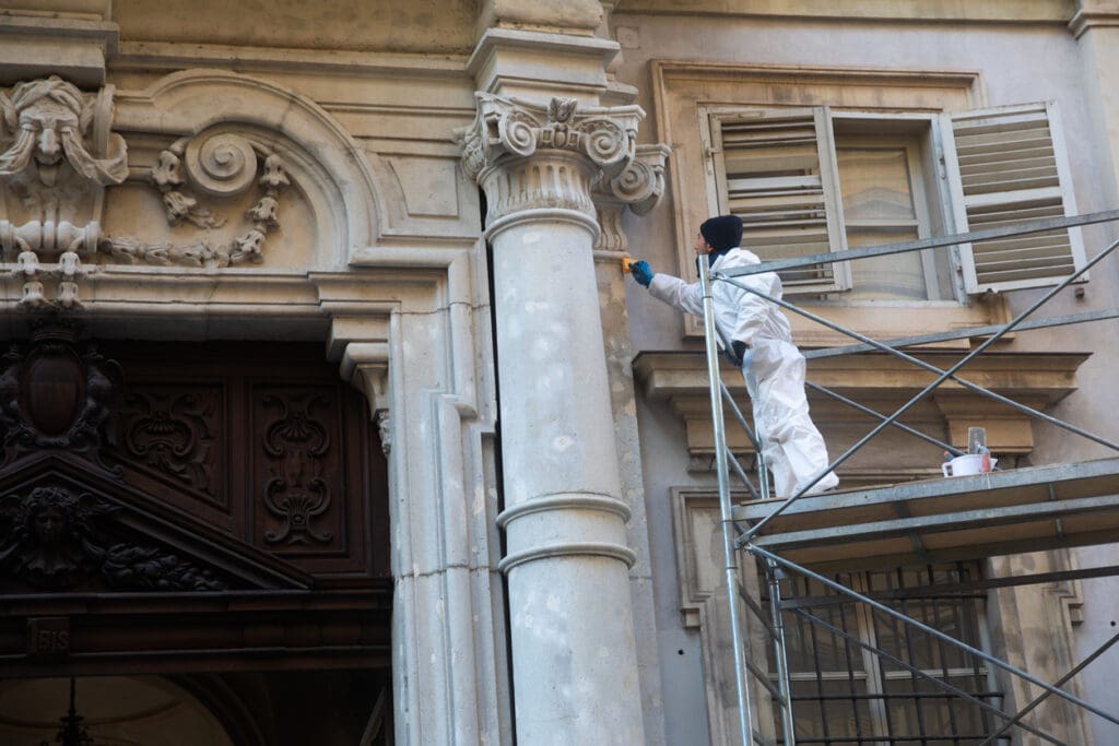 A worker in white protective clothing stands on scaffolding, painting or restoring the ornate exterior of an old stone building with columns and decorative carvings. An open window is visible nearby.