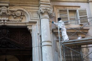 A worker in white protective clothing stands on scaffolding, painting or restoring the ornate exterior of an old stone building with columns and decorative carvings. An open window is visible nearby.