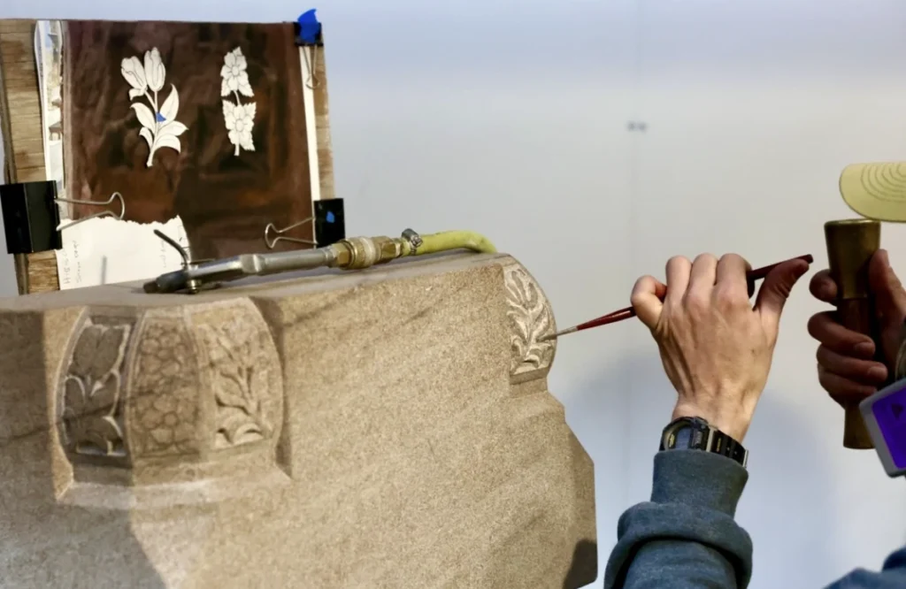 A person uses a chisel and mallet to carve floral designs into a stone block, with reference sketches of flowers clipped nearby for guidance.