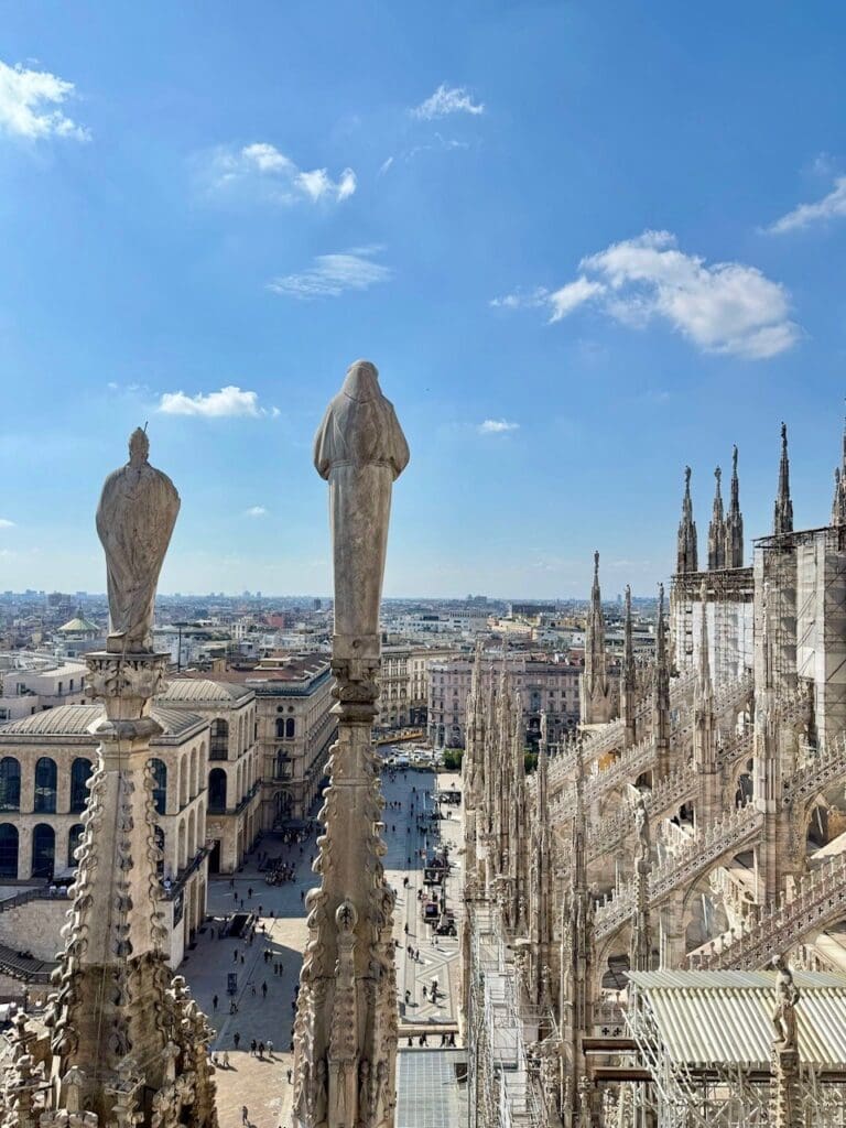 View from the rooftop of Milan Cathedral, built with stone from Candoglia Quarry, showing detailed statues and spires overlooking Piazza del Duomo with people below under a blue sky dotted with clouds.