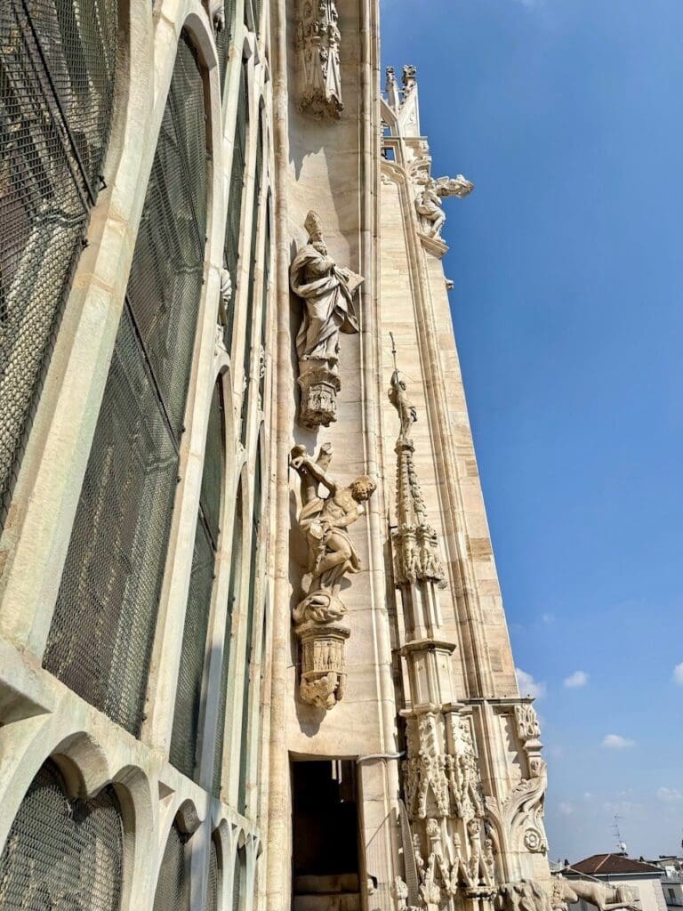 A close-up view of ornate statues and intricate architectural details on the exterior of Milan Cathedral, built from Candoglia Quarry stone, under a clear blue sky.