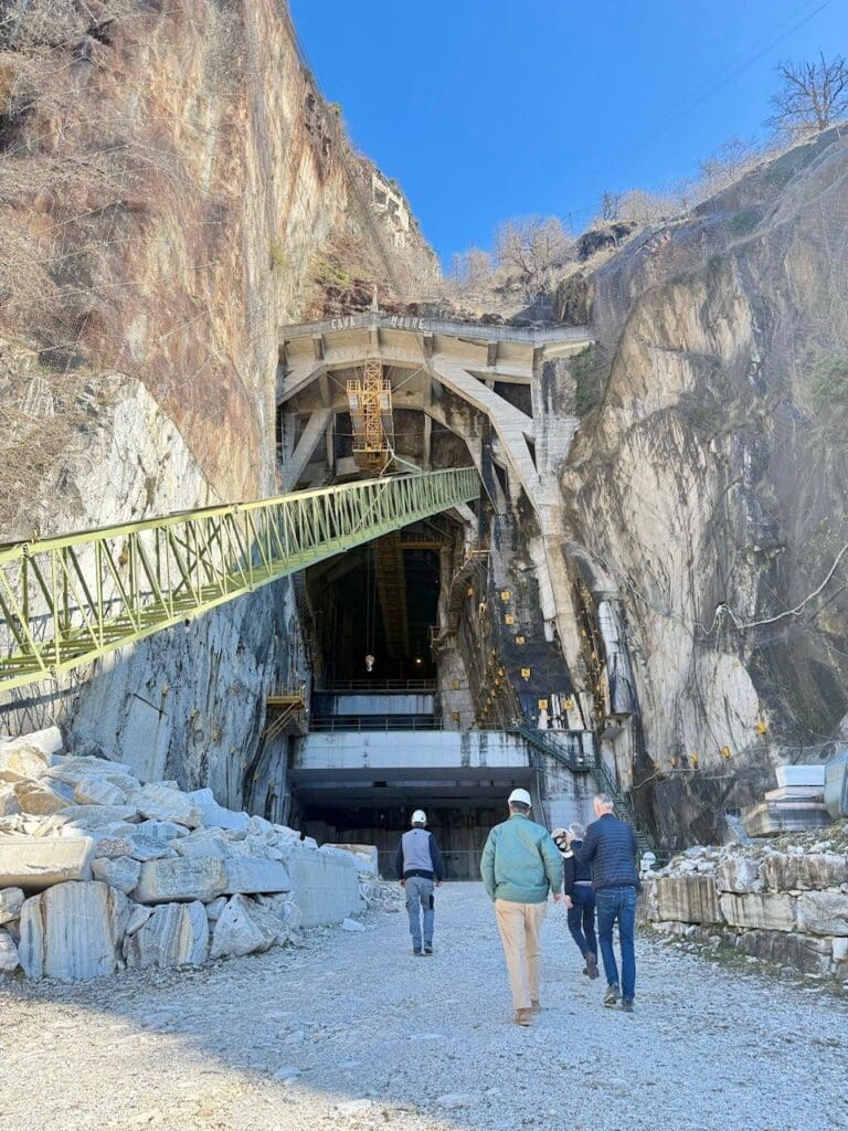 Four people walk toward a large marble cave entrance at the Candoglia Quarry, where marble for Milan Cathedral was sourced. A metal bridge extends above them, piles of marble blocks lie nearby, all beneath a clear blue sky.