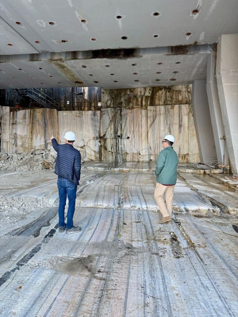 Two people wearing hard hats stand in the Candoglia Quarry, where stone for the Milan Cathedral was sourced, examining the exposed rock walls and floor, with construction debris scattered around the area.