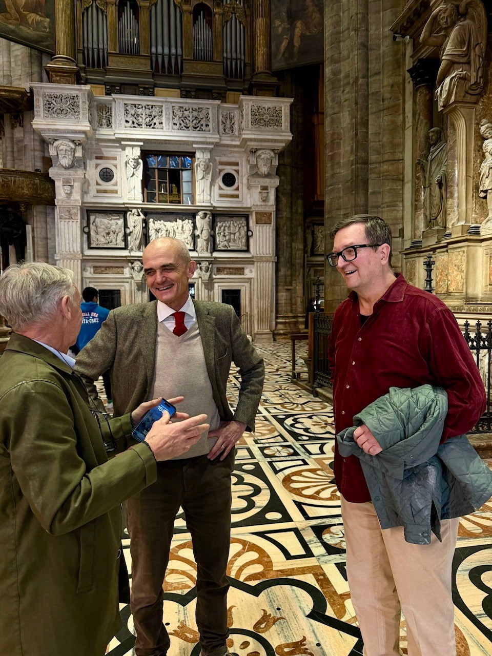 Three men stand talking inside an ornate, historic building with intricate patterned floors and detailed stone carvings, reminiscent of Milan Cathedral’s grandeur. One man gestures as the others smile, their conversation echoing tales from the Candoglia Quarry.