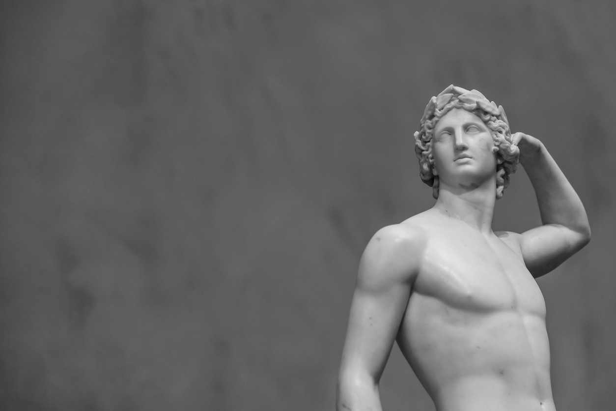 Black-and-white photo of a classical marble statue of a nude male figure with curly hair, standing with one arm bent behind his head, gazing upward, against a plain background.