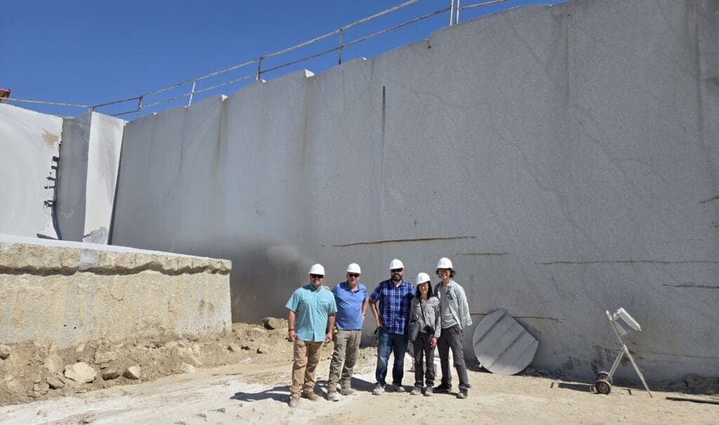 Five people wearing hard hats and casual clothing stand together at the Sierra White Quarry, their figures framed by tall, smooth stone walls and construction materials under a clear blue sky.