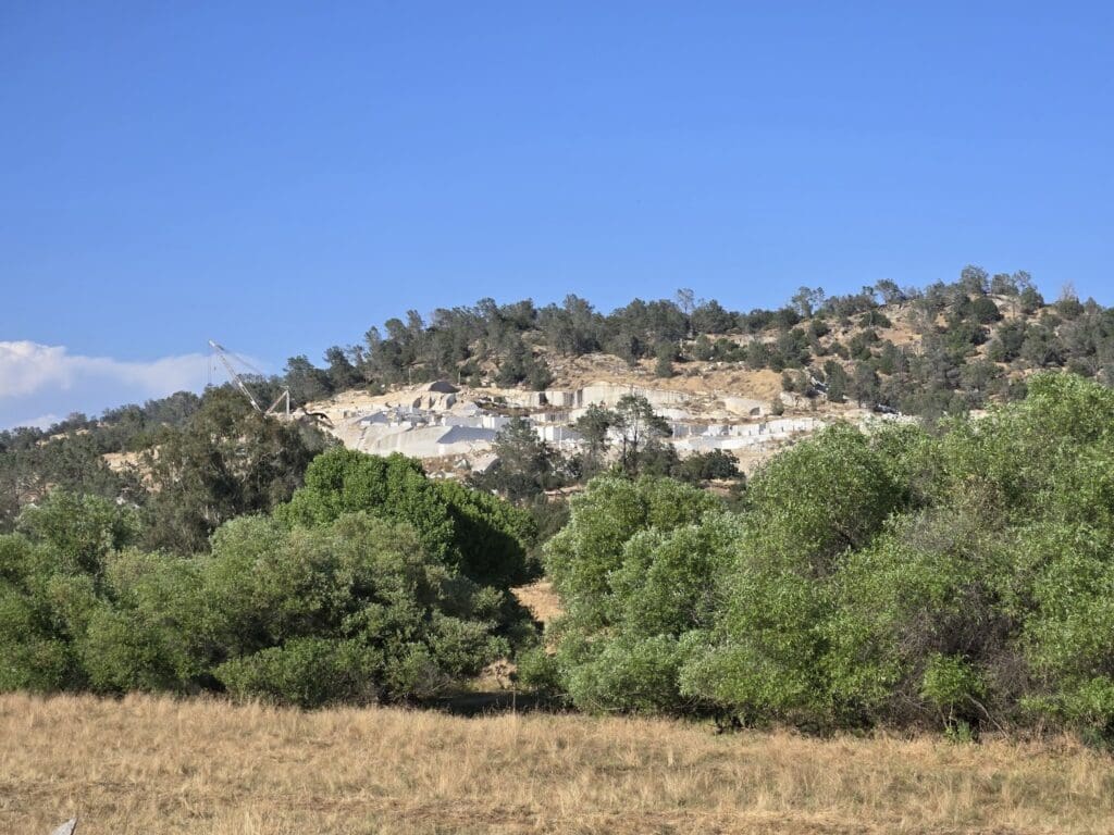 A hillside quarry, known as the Sierra White Quarry, features exposed white rock and machinery in the background, surrounded by trees and green vegetation under a clear blue sky.