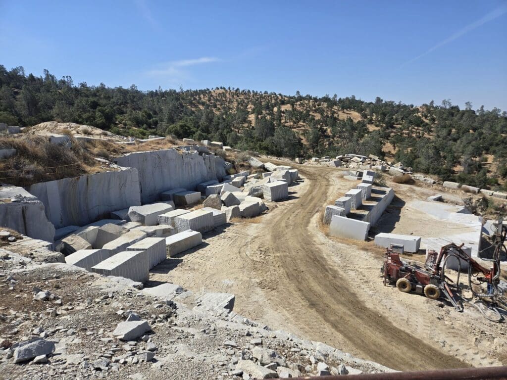 A large outdoor stone quarry with rectangular blocks of cut Quarra Stone arranged along a dirt road; machinery and scattered debris are visible, with trees and hills in the background under a clear blue sky.