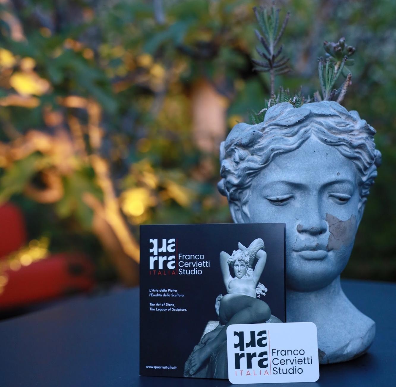 A stone bust of a womans head with cracks is displayed outdoors on a table next to a Franco Cervietti Studio brochure and card, set against softly blurred greenery at the Annual Gathering in Pietrasanta, Italy.