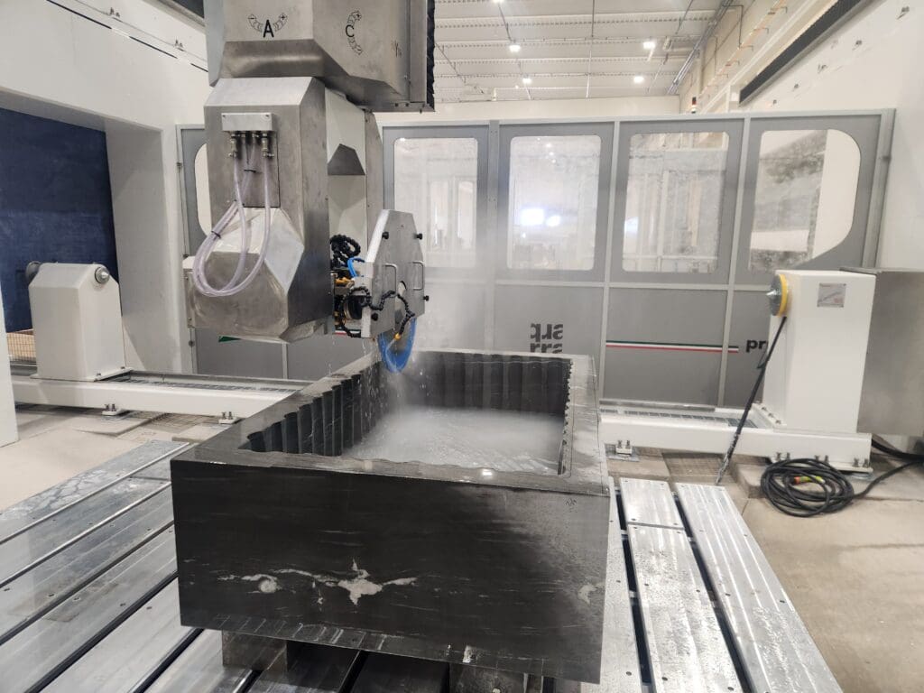 A large industrial machine from Quarra Stone uses a water-cooled circular saw to cut a rectangular stone block in a modern, well-lit factory setting. Water spray and mist are visible around the cutting area, reminiscent of scenes in Bloomberg Businessweek.