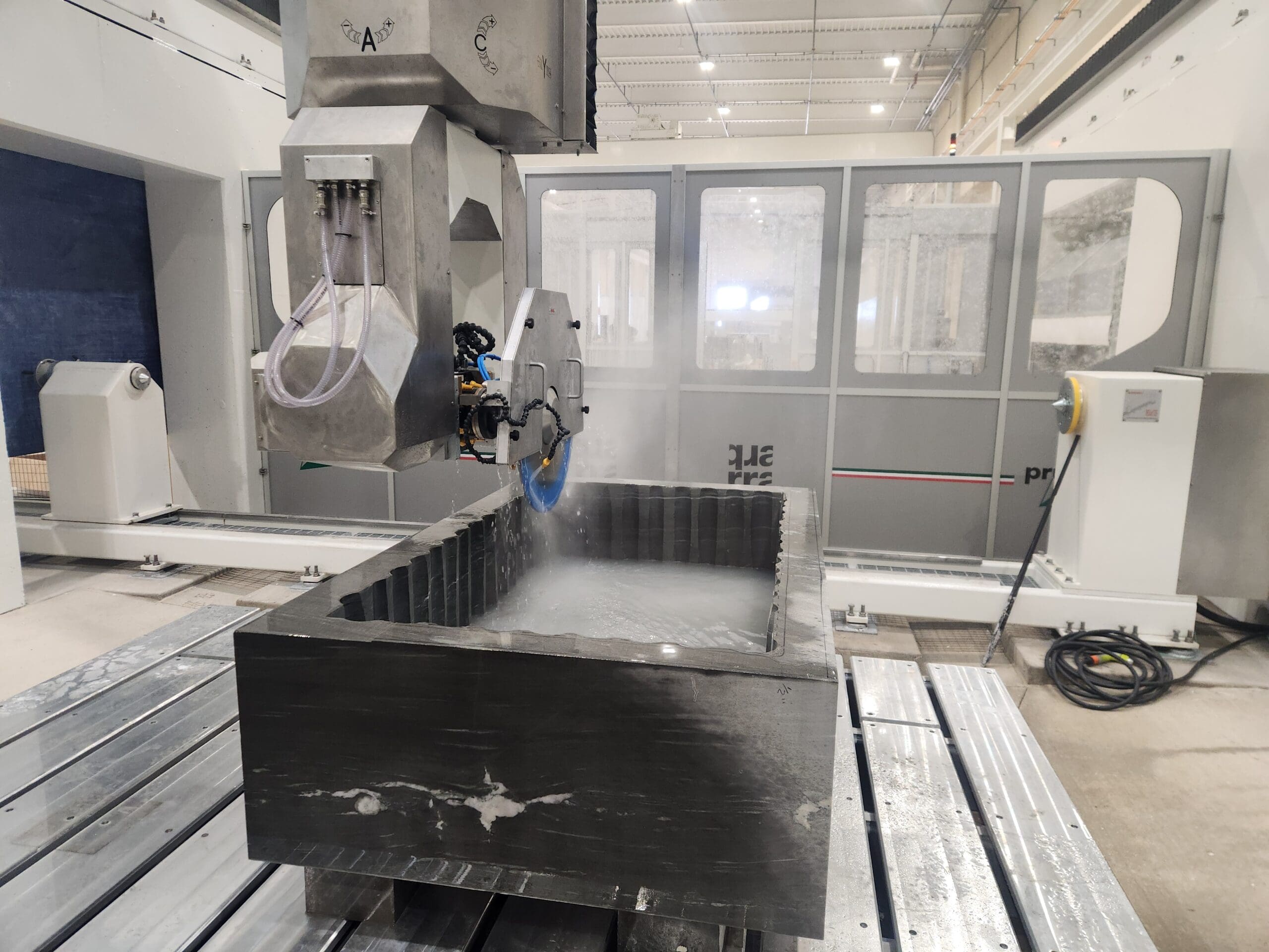 A large industrial machine from Quarra Stone uses a water-cooled circular saw to cut a rectangular stone block in a modern, well-lit factory setting. Water spray and mist are visible around the cutting area, reminiscent of scenes in Bloomberg Businessweek.