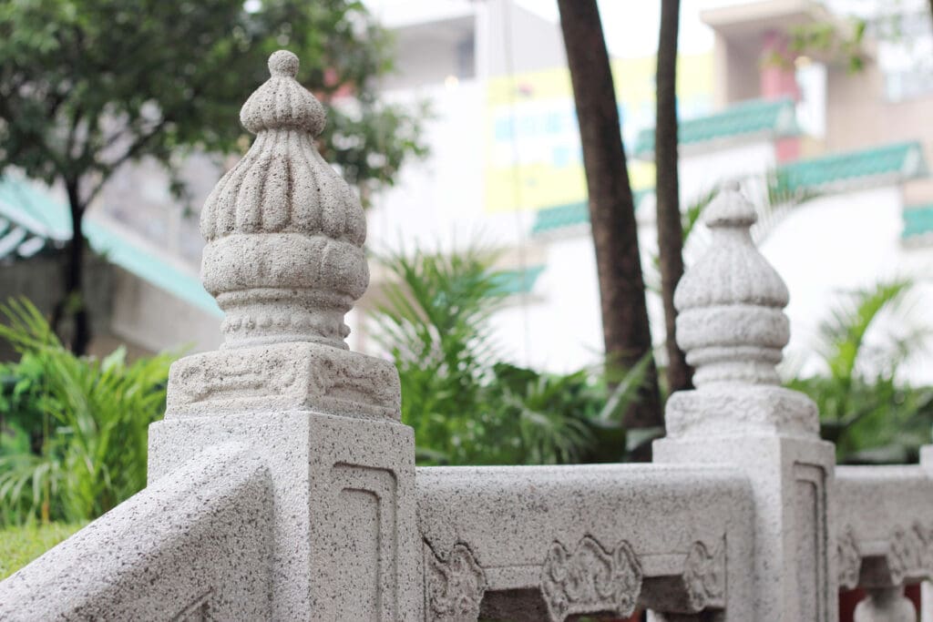Close-up of two ornate architectural stone balusters on a decorative railing in a garden setting, with green foliage and blurred buildings in the background.