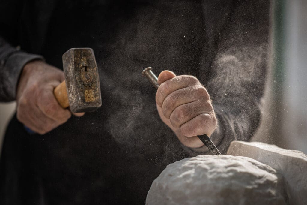 Close-up of a person’s hands carving architectural stone with a chisel and hammer, creating dust as they work. The background is blurred, focusing on the detailed craftsmanship and motion.