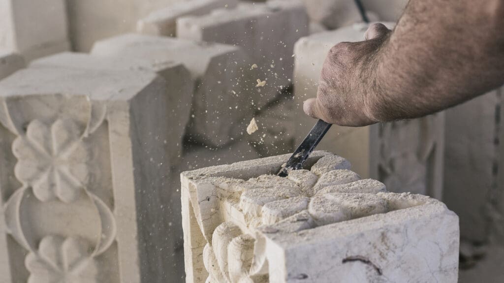 A close-up of a person’s hand using a chisel to carve intricate floral patterns into hand-finished stone, with small stone chips flying in the air.