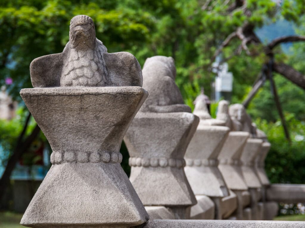 A row of stone sculptures featuring falcon-like bird figures perched on hourglass-shaped bases, crafted from Vals Quartzite, set outdoors with green trees in the background.