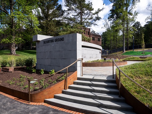 A curved stone Burying Ground Memorial stands beside a pathway and landscaped area with stairs and metal railings, surrounded by trees and greenery under a partly cloudy sky.