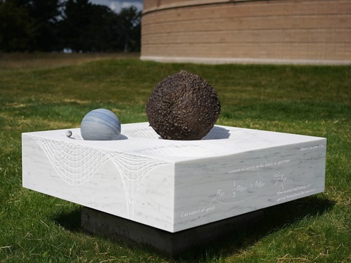 A sculpture titled Time Will Tell features a textured dark sphere and a smooth blue-gray sphere on a white marble base, engraved with wave patterns and scientific notations, displayed outdoors on grass.