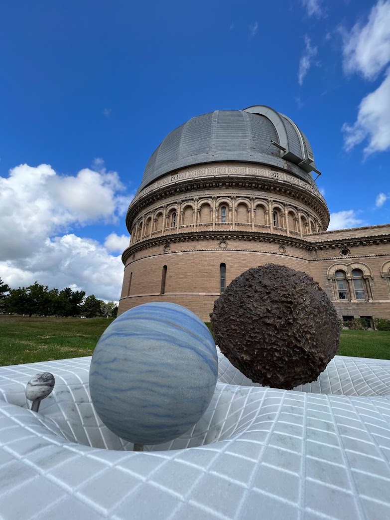 A large observatory with a domed roof stands in the background. In the foreground, the Time Will Tell sculpture displays models of planets and a textured black sphere on a grid, illustrating gravitational distortion. The sky is partly cloudy and blue.