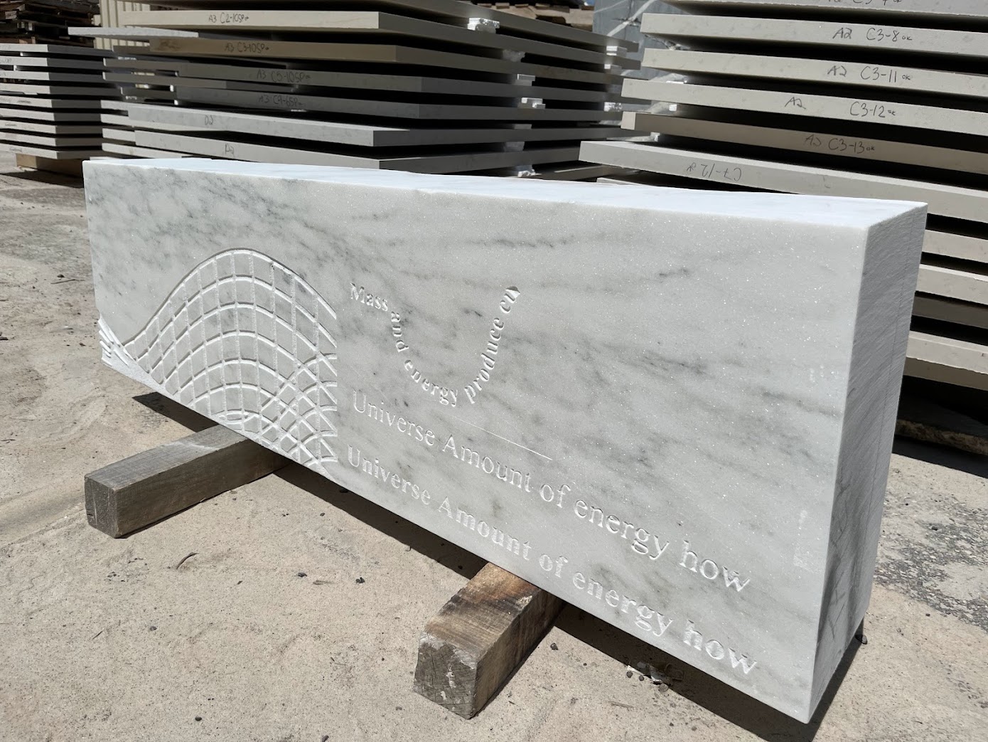 A large rectangular slab of white marble with engraved text and a curved wave pattern sits outdoors on wooden supports, surrounded by stacks of similar stone slabs—its inscription reads, “Time Will Tell.”.