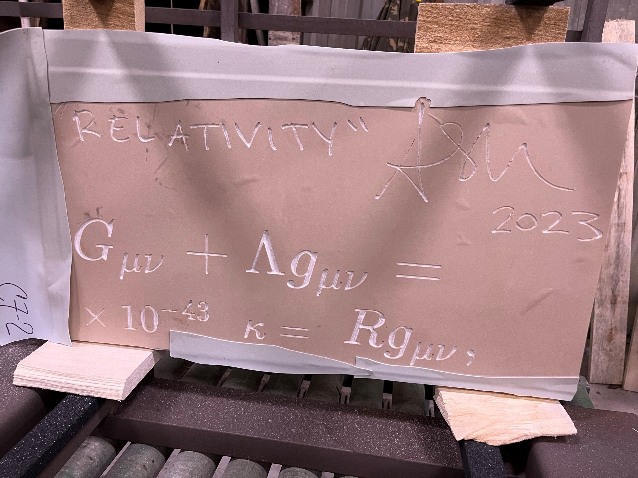 A clay tablet displays the word RELATIVITY, an autograph, 2023, and Einstein’s field equation—Gμν + Λgμν = (8πG/c⁴)Tμν—inscribed in white. The phrase “Time Will Tell” is delicately etched below, as the tablet rests on wooden supports.