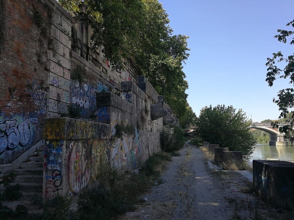 A stone path, maintained with the best stone preservation practices for long-term longevity, runs alongside a river, bordered by a tall, graffiti-covered wall and overhanging trees. In the distance, a bridge spans the river under a clear blue sky.