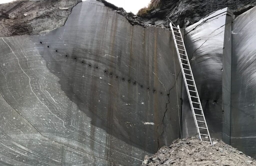 A tall metal ladder leans against a steep, smooth quartzite rock face with visible striations and drilled holes. The natural stone surface is dark gray, and loose gravel sits at the base of the wall.