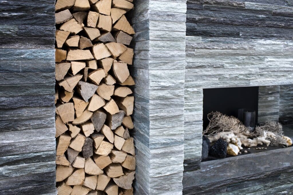 Stacked firewood neatly arranged in a vertical alcove beside a modern architectural stone fireplace, which contains decorative branches and stones. The fireplace has a sleek, contemporary stone finish.