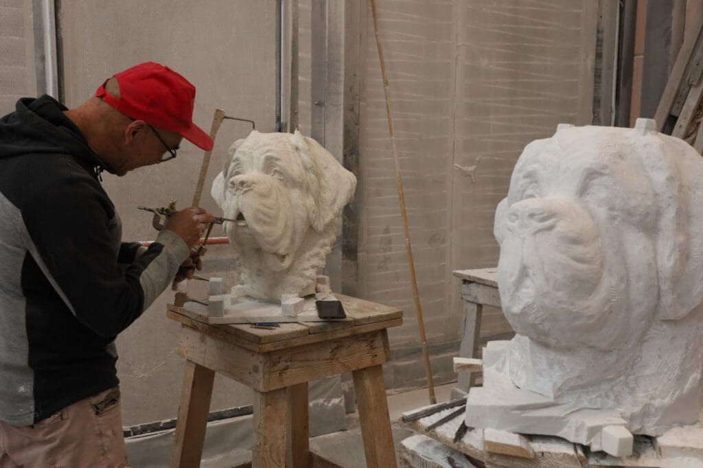A man in a red cap sculpts a large dog head out of Swiss stone on a wooden stand, with another similar dog sculpture nearby in an artist’s studio.