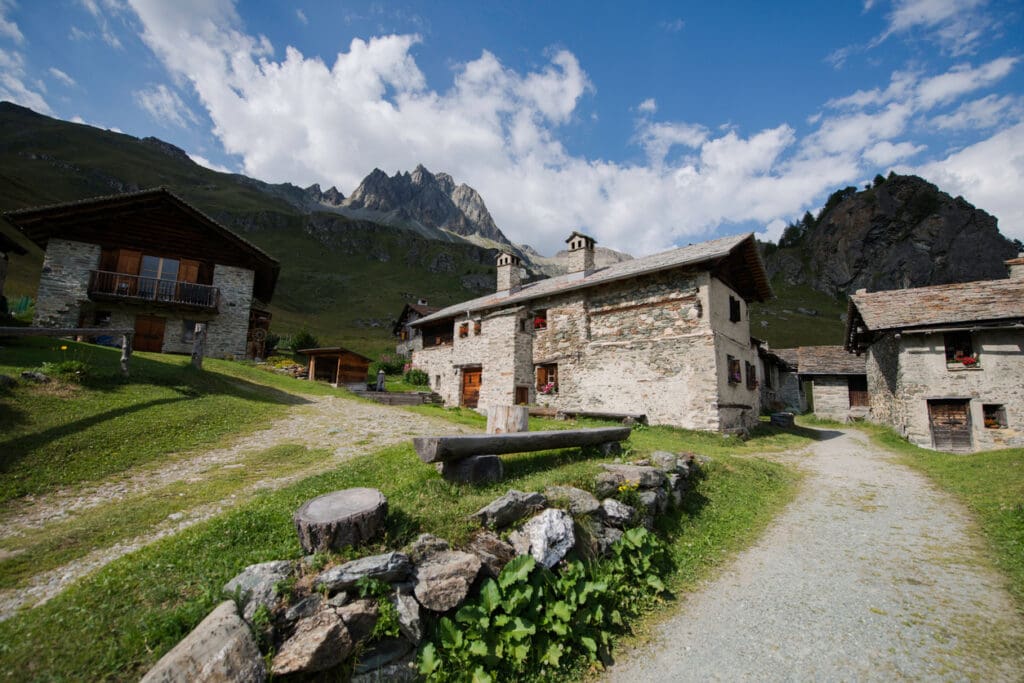 Stone houses built of natural stone with wooden roofs sit on a grassy hillside, surrounded by mountains under a blue sky. A dirt path and rustic benches in the foreground complete this serene rural scene inspired by Peter Zumthor’s timeless style.