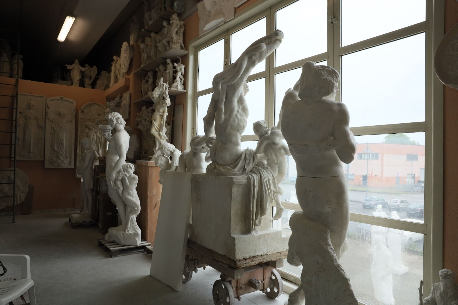 Several white marble statues, including classical human figures and busts, are displayed in a studio with large windows. Natural light highlights each marble sculpture, hinting at the intricate process from quarry to finished art. A building and parked cars are visible outside.