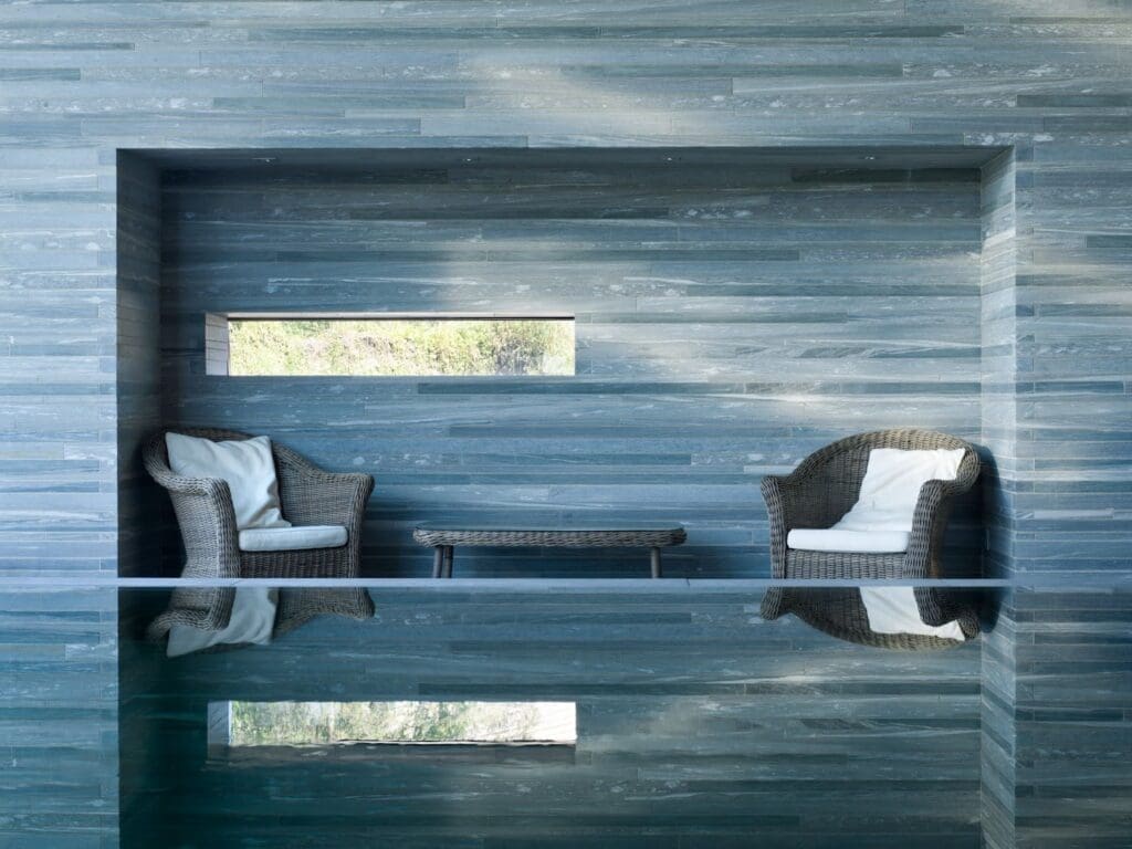 Two wicker chairs with white cushions and a low table sit against a sleek gray Vals Quartzite wall with a rectangular window, reflected in the calm indoor pool—a stylish nod to modern architecture.