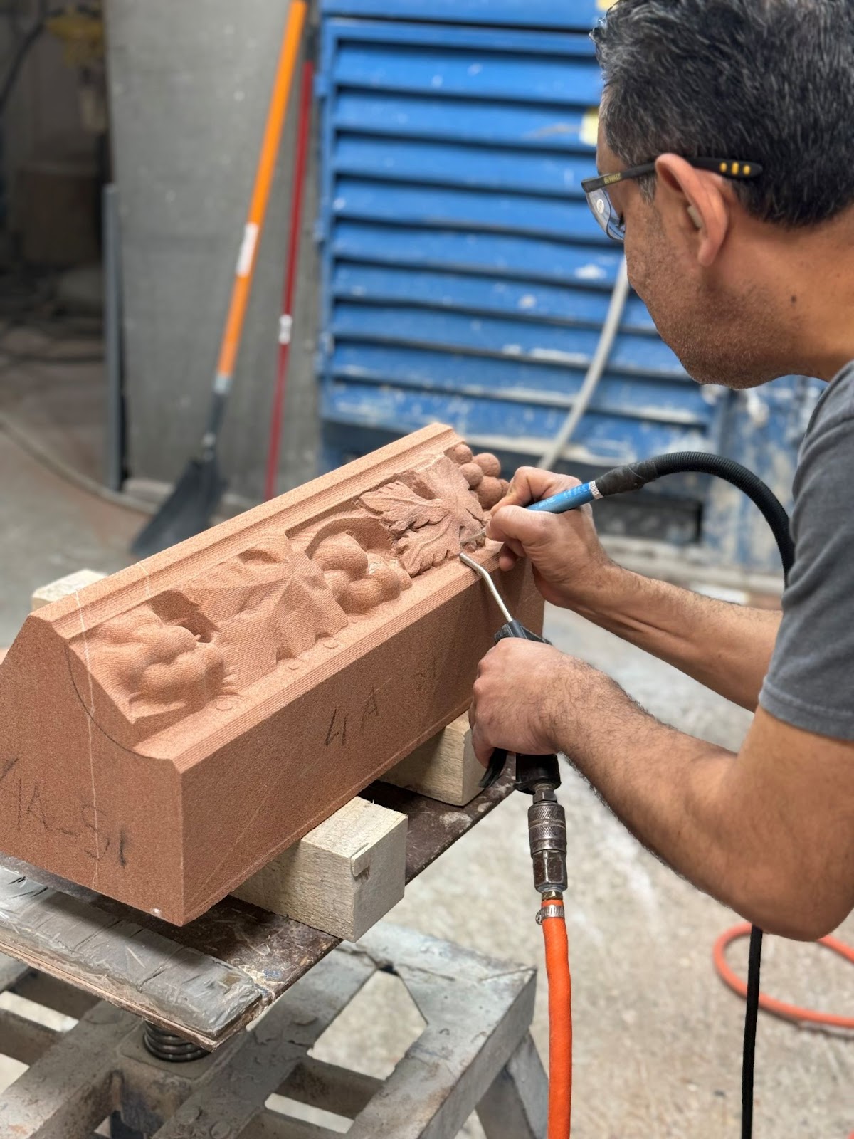 A man uses a power tool to carve detailed designs into a reddish-brown stone block in a workshop, blending traditional stone fabrication with technology; blue metal sheets and tools are visible in the background.