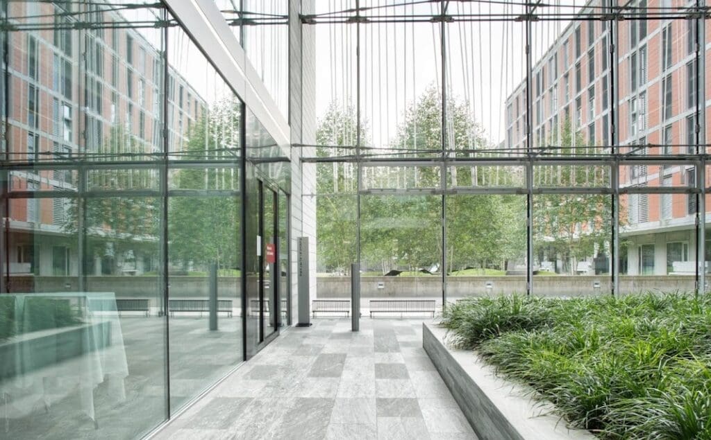 A modern glass building interior with large windows, gray tiled floors of architectural stone, green indoor plants, and a view of trees and red-brick buildings outside. Natural light fills the space.