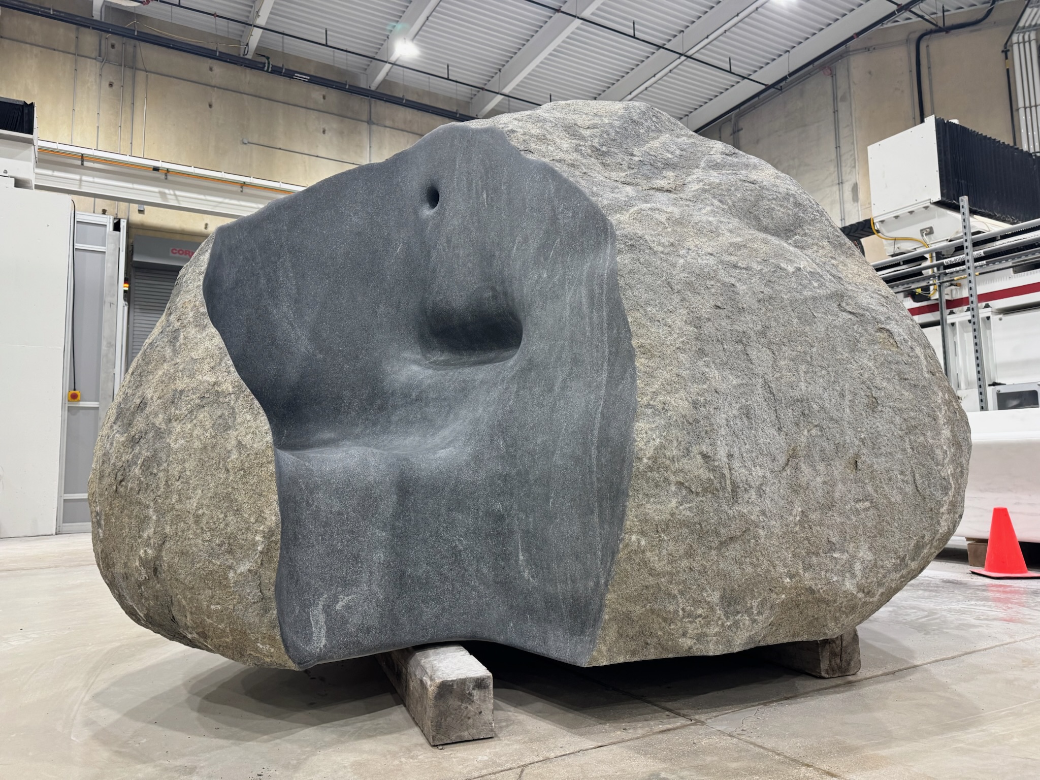 A large, irregularly shaped boulder sits indoors on wooden blocks. Part of its surface is smoothly carved into a seat and backrest, as if inviting you to pause and listen—Whispering Through a Stone. Industrial equipment and a traffic cone are visible in the background.
