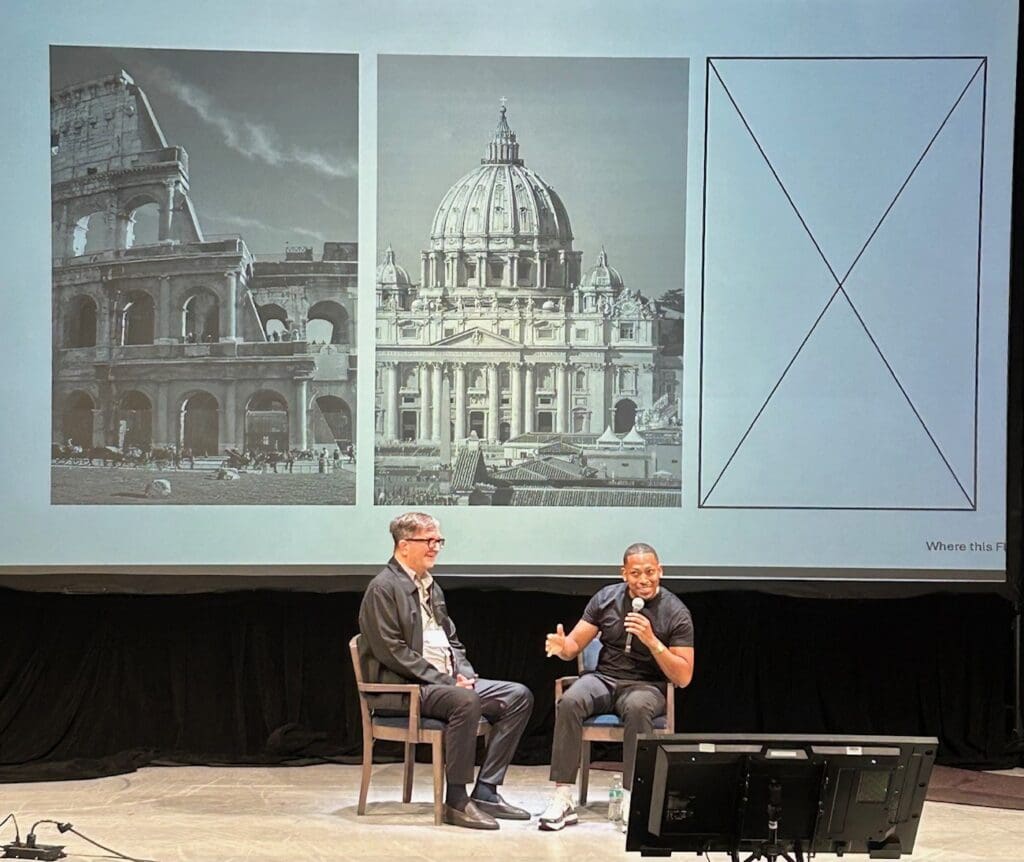 Two men sit on a stage in front of a screen showing black-and-white images of the Colosseum, St. Peter’s Basilica, and an empty box—one gesturing as he speaks about Quarra Stone, while the other listens attentively.