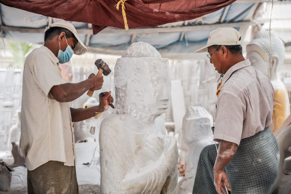 Two men work on sculpting a large white architectural stone statue of a seated figure; one uses a hammer and chisel while the other observes. Both wear hats and traditional attire, and one wears a face mask.