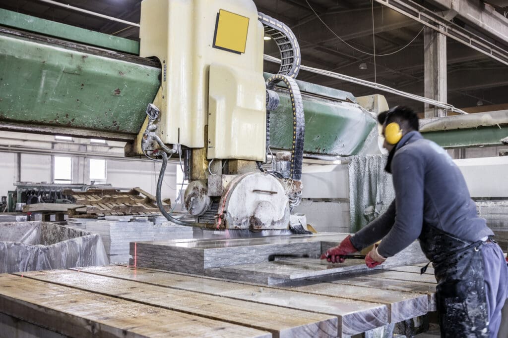 A worker wearing ear protection operates a large industrial stone cutting machine in a factory, precisely cutting slabs of Vals Quartzite on a wooden work surface.