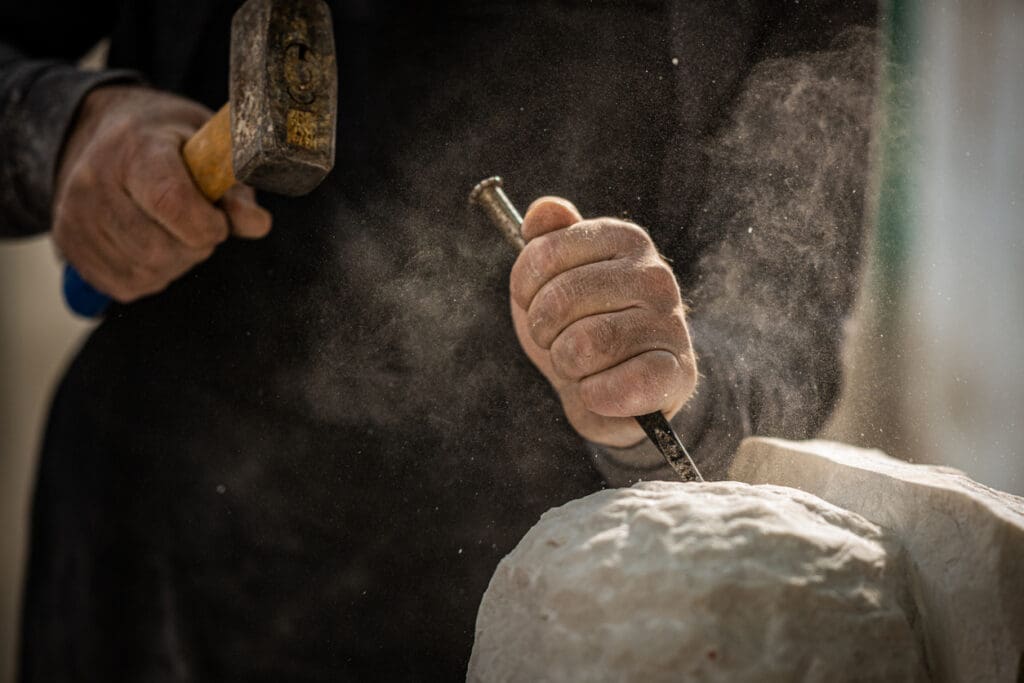Close-up of a person’s hands carving stone with a chisel and hammer, dust swirling in the air—a craftsman in dark clothing focusing intently on shaping the rough surface for custom commercial stonework.