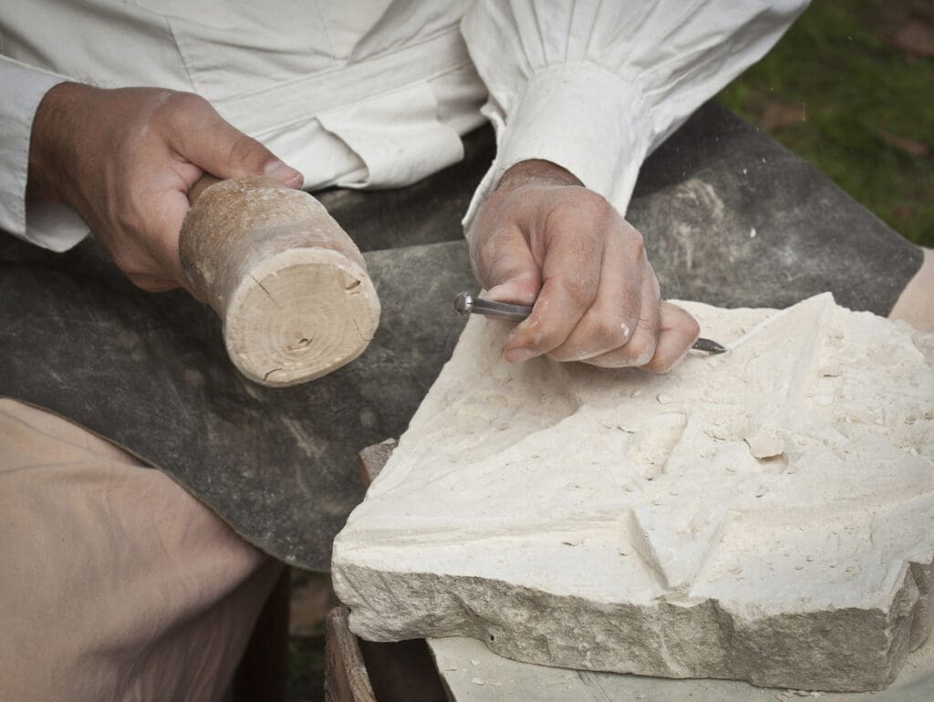 A person wearing a white shirt carves a design into a quartzite block using a chisel and wooden mallet, with stone chips scattered on the surface.
