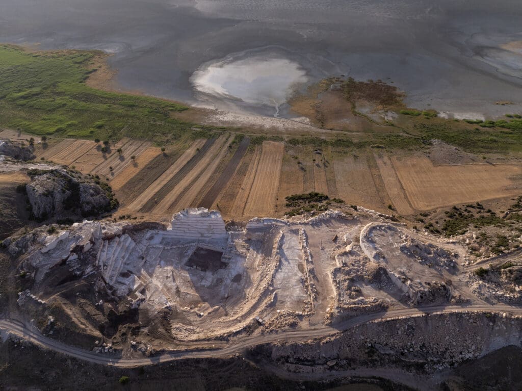 Aerial view of a limestone quarry with terraced excavation, adjacent to farmland with green and brown fields, and a salt flat or dried lakebed in the background—ideal for sourcing architectural stone like quartzite favored by Peter Zumthor.