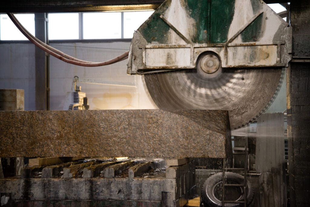 A large circular saw cuts through a thick slab of Vals Quartzite in an industrial workshop, with water spraying to cool the blade—evoking the precision found in Peter Zumthor’s use of architectural stone.