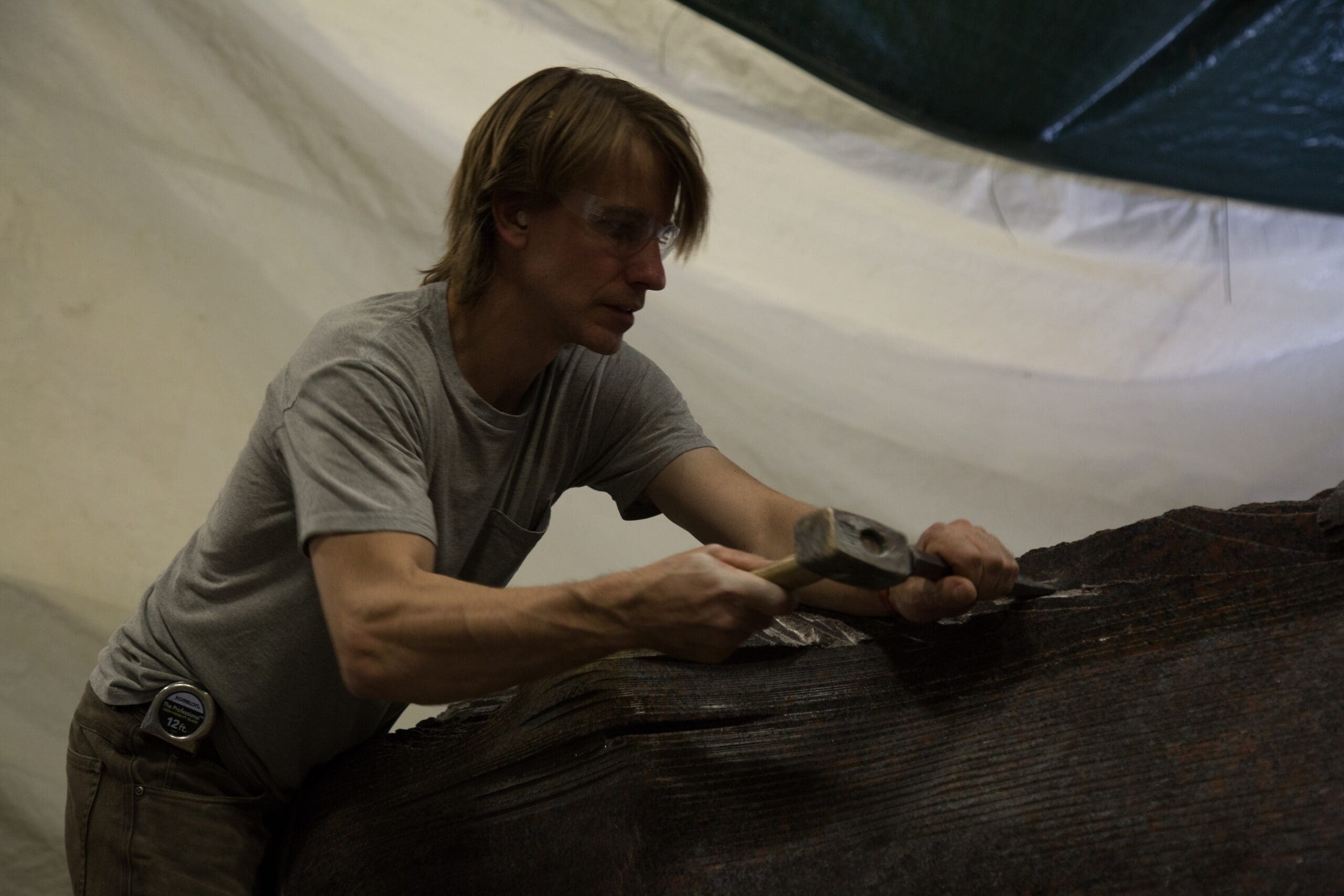 Miss Madison, wearing safety glasses, uses a hammer and chisel to work on a large, dark stone surface under a white and green tarp.