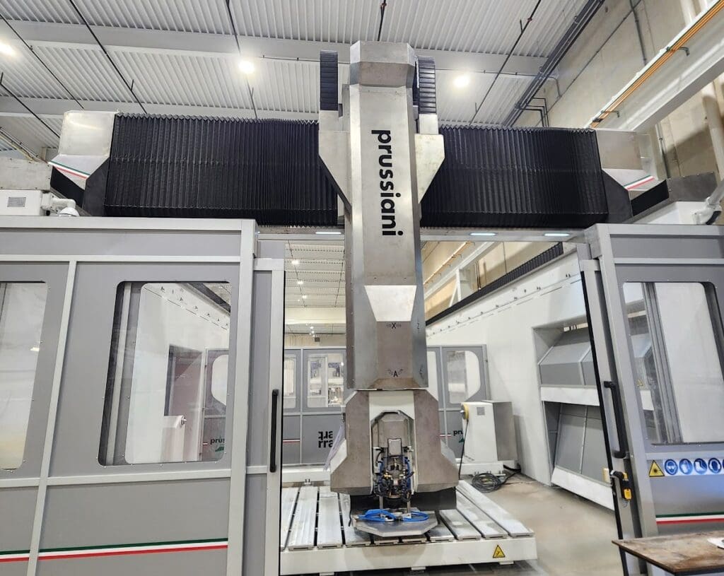 A large industrial CNC machine from Prussiani stands in a factory setting, with metallic parts, glass-paneled enclosures, and overhead lights visible in the background.