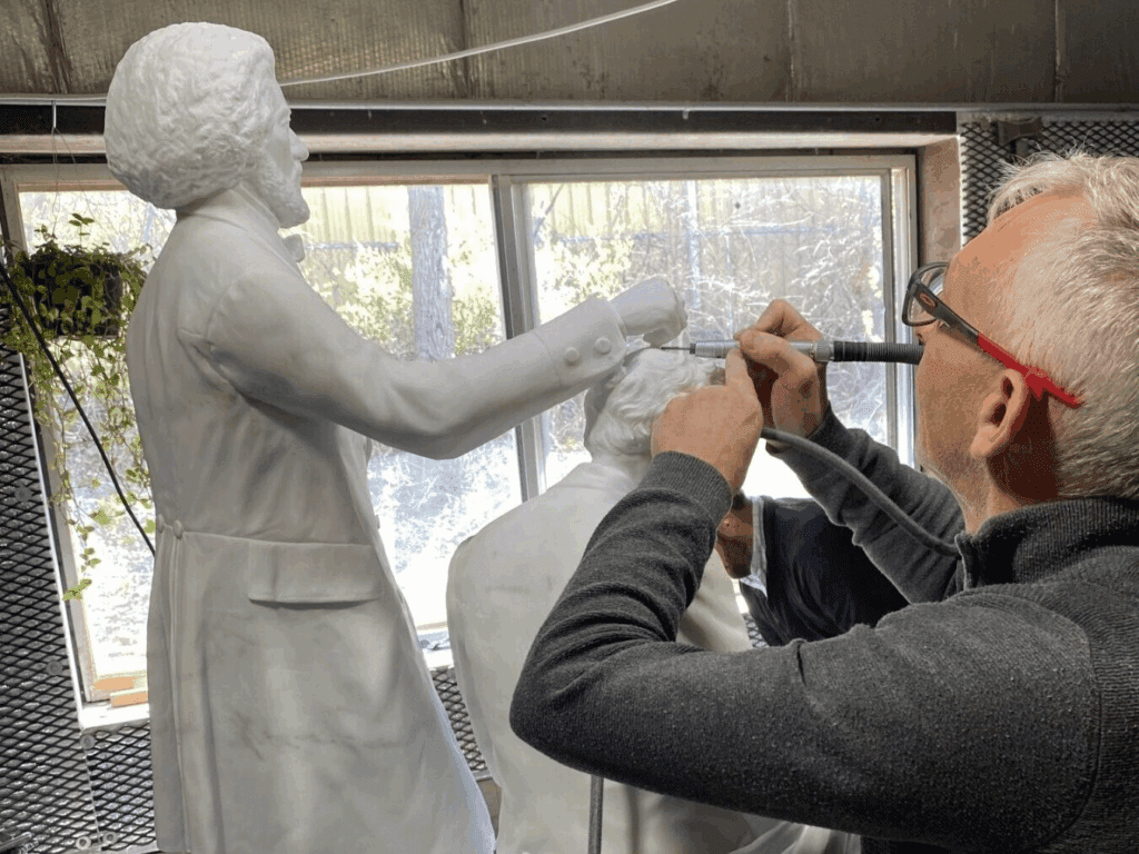 A sculptor with gray hair and glasses uses a tool to work on a white Swiss stone statue of two standing figures in a well-lit studio with large windows.