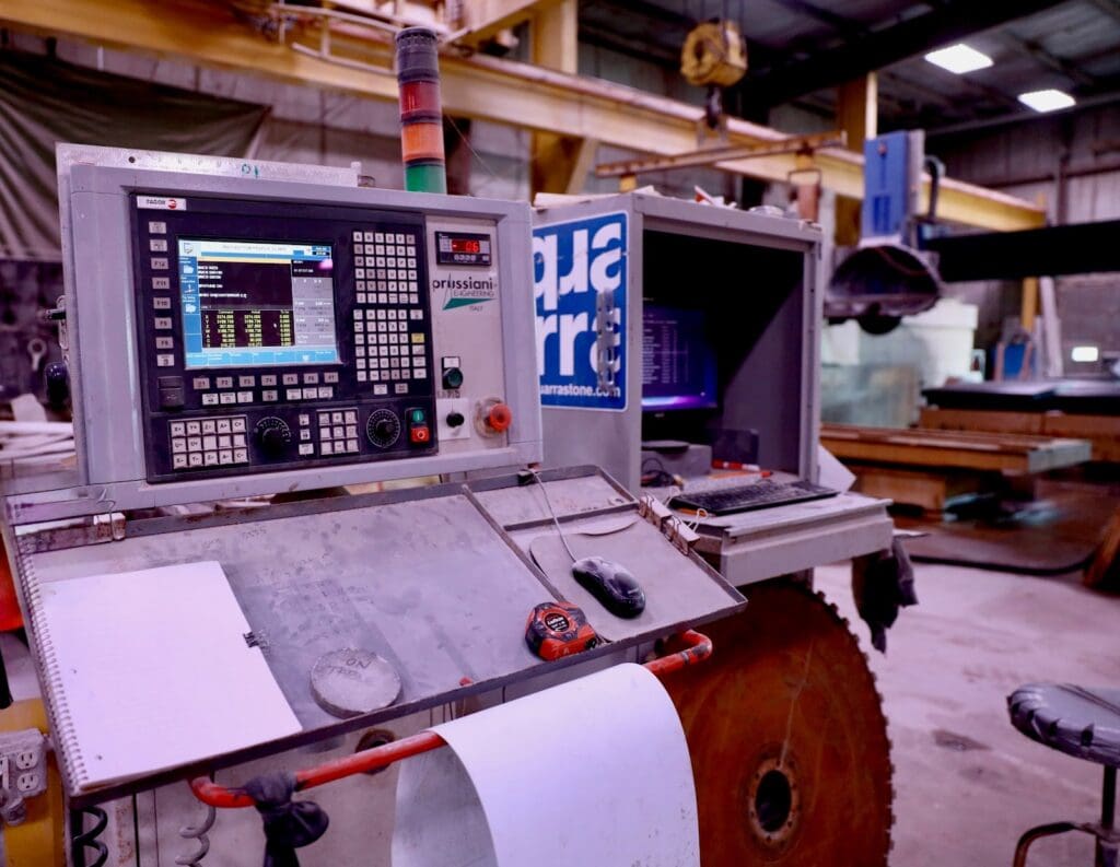 A close-up of a CNC machine control panel with multiple screens and buttons, set in an industrial workshop where large machinery shapes natural stone like exquisite Vals Quartzite, echoing the precision admired by architects such as Peter Zumthor.