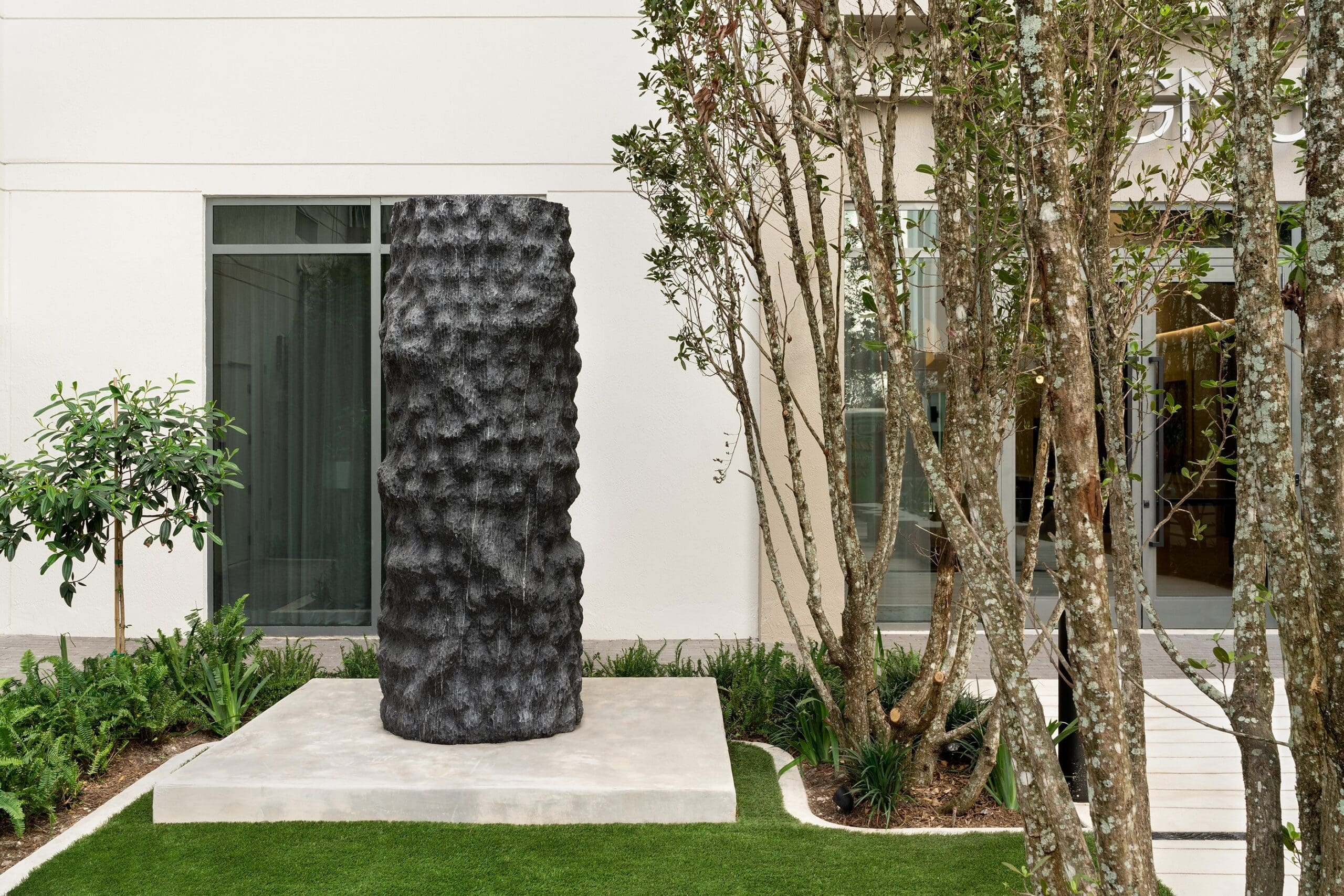 A textured, cylindrical black sculpture stands on a concrete platform in a landscaped courtyard, reflecting the enduring influence of ancient architecture amid green grass, trees, and a modern building with large windows in the background.