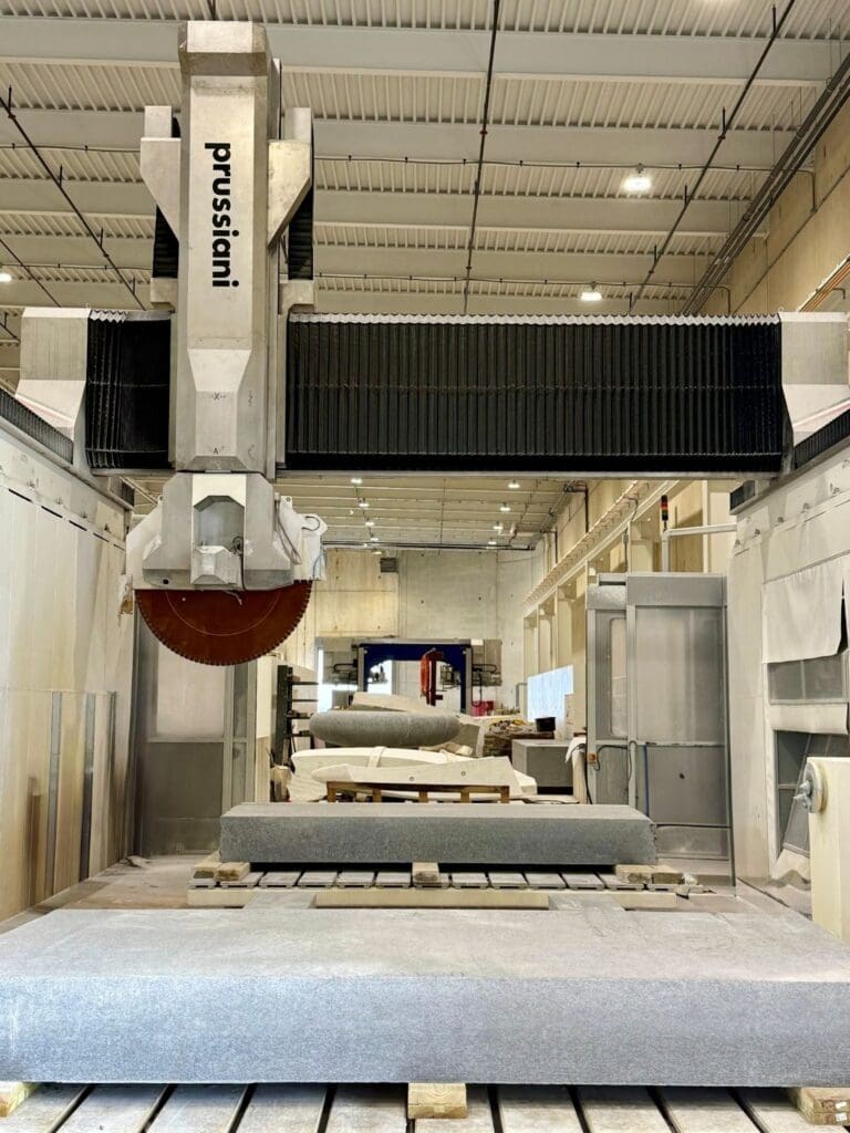 A large industrial stone-cutting machine labeled Prussiani in a spacious, modern factory, with a slab of stone positioned underneath the cutting blade.