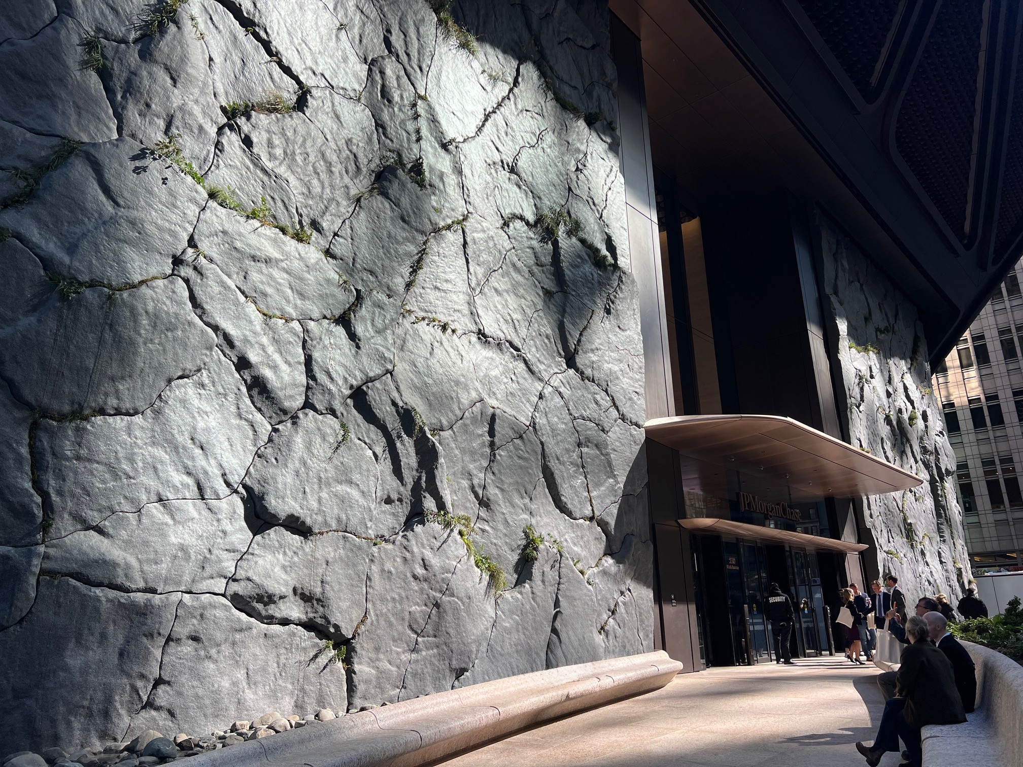 Modern building facade designed to look like cracked stone, echoing the organic forms in Maya Lin A Parallel Nature, with plants growing in crevices; several people stand and sit near the entrance in sunlight and shade.